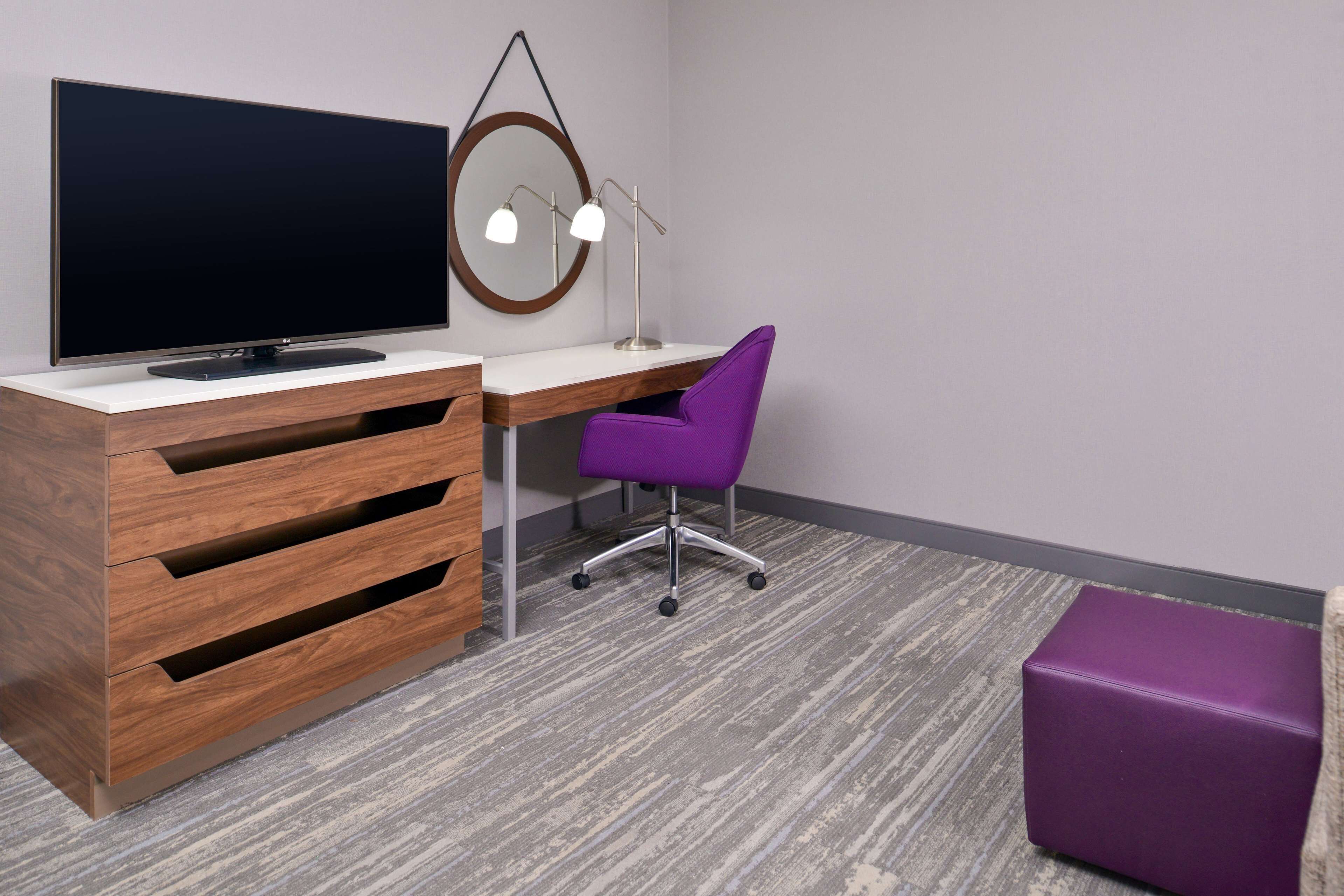 Un espacio de oficina moderno con un escritorio de madera, una silla de oficina púrpura, un televisor y un espejo redondo con luces.