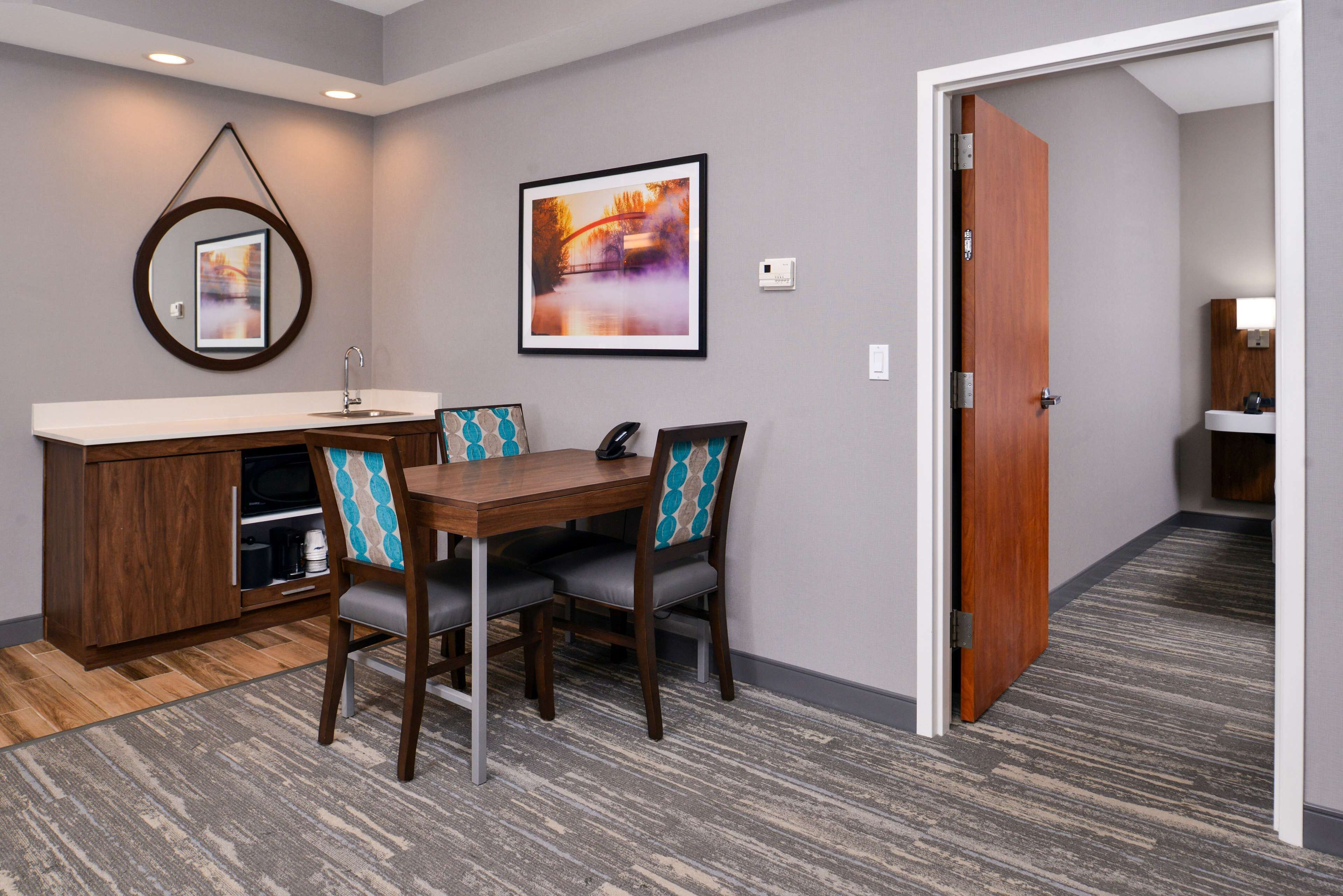 Habitación de hotel con comedor, que incluye una mesa y sillas de madera, una encimera de cocina integrada y un cuadro grande en la pared.