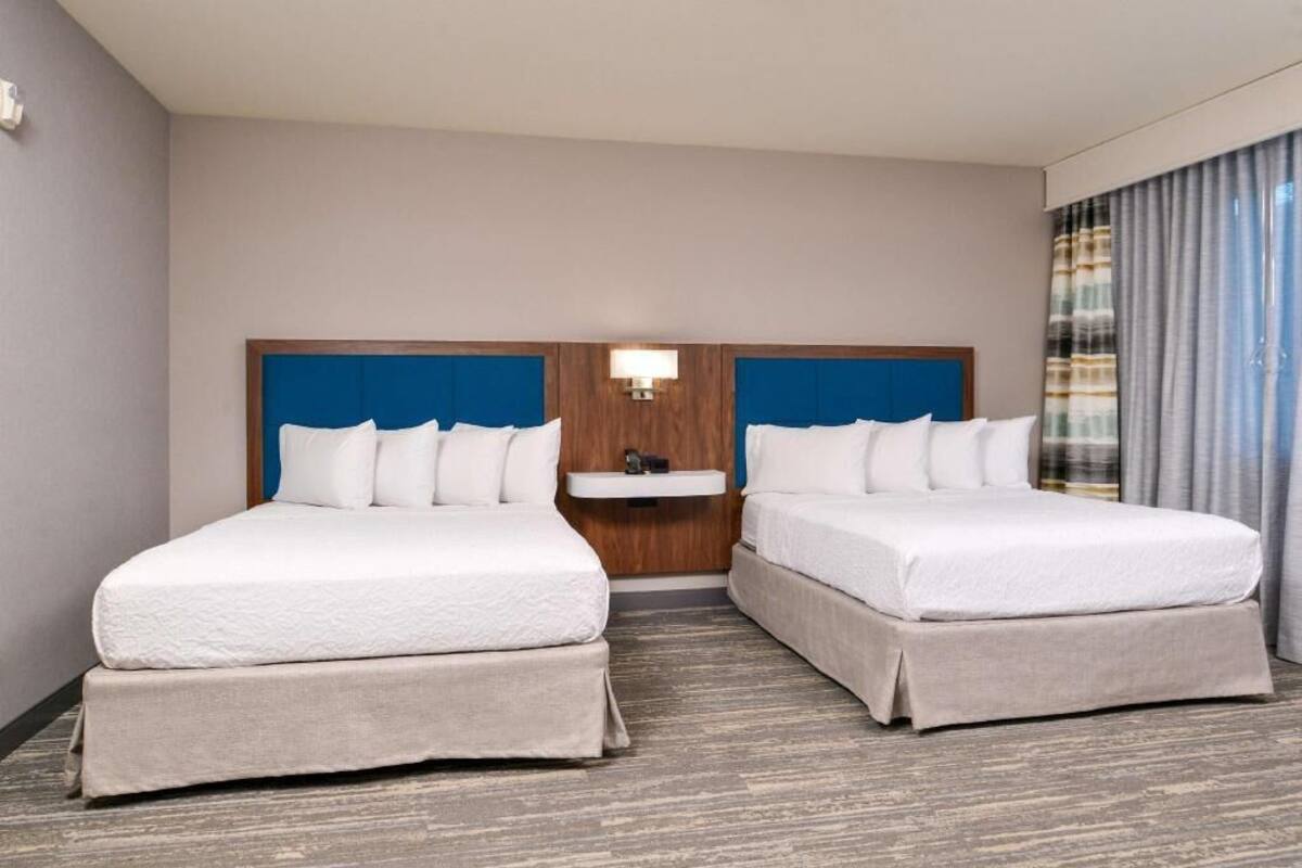 A hotel room with two beds, a small bedside table, a wall-mounted light, and a window with curtains.