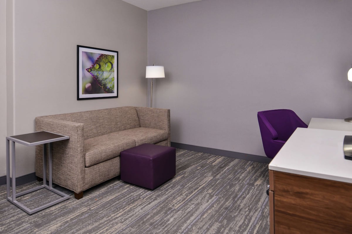 Habitación de hotel con alfombra gris, sofá gris, reposapiés morado, mesita auxiliar pequeña, silla morada, escritorio blanco y un cuadro en la pared.