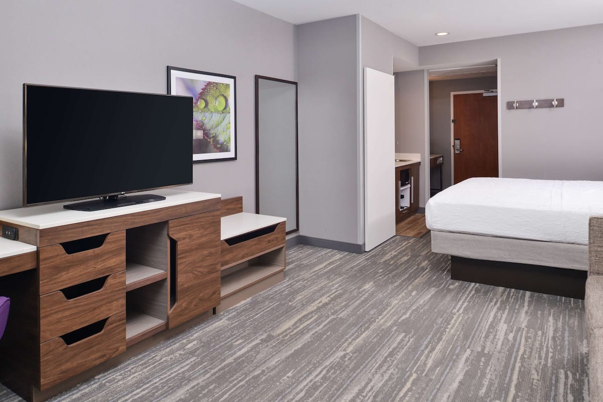 Una habitación de hotel moderna con televisor de pantalla plana, una cama, un escritorio con cajones y una puerta que da a otra habitación.