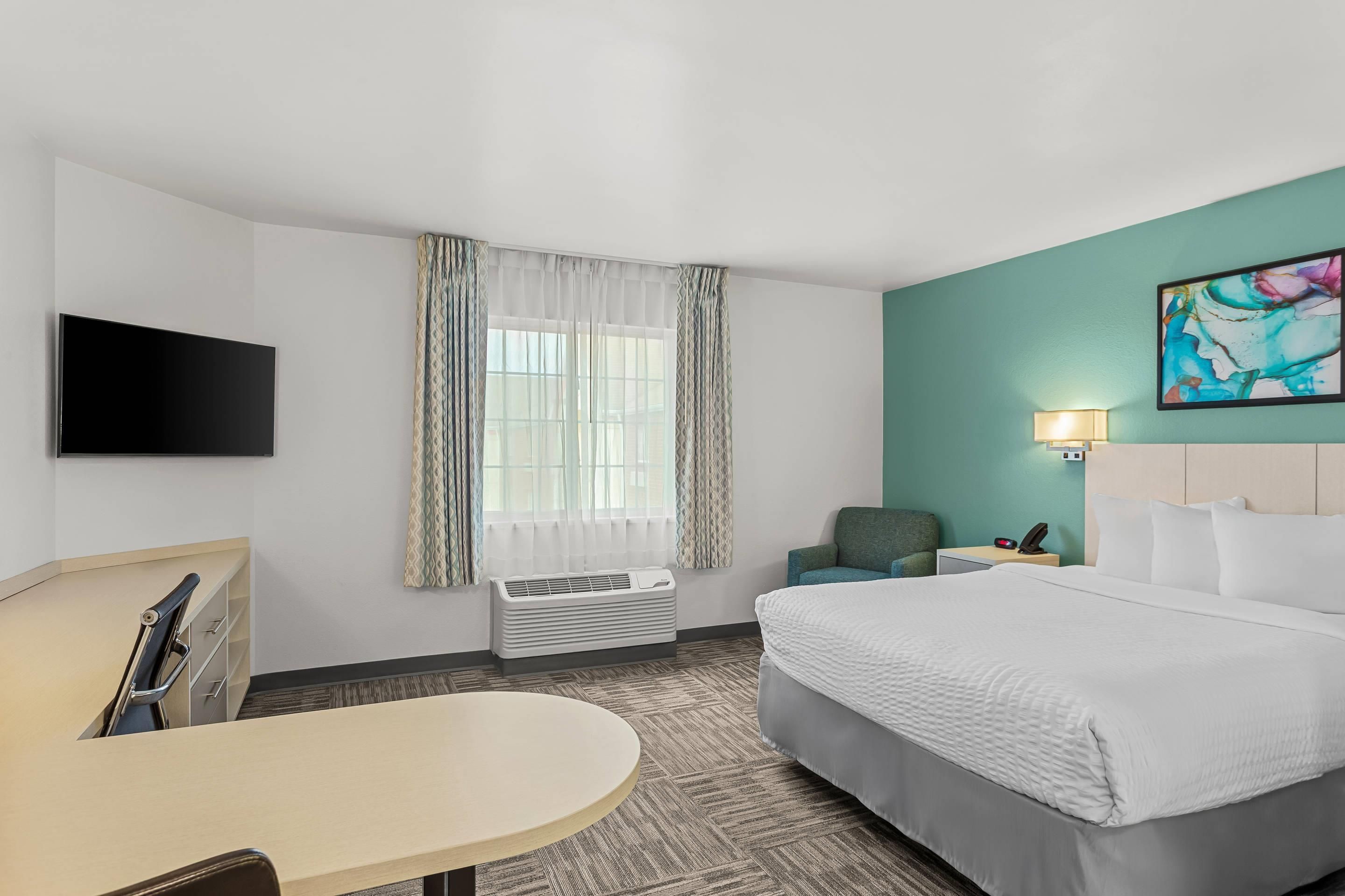 Una habitación de hotel con cama, escritorio, silla, televisor, ventana con cortinas, un cuadro y una lámpara de noche.
