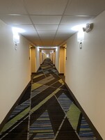 Un pasillo de hotel con alfombra estampada, luces fijadas a la pared y puertas a ambos lados.