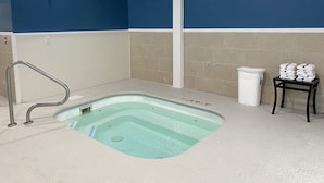 A small, rectangular hot tub with a metal handrail, beige tiled walls, and a white trash can.