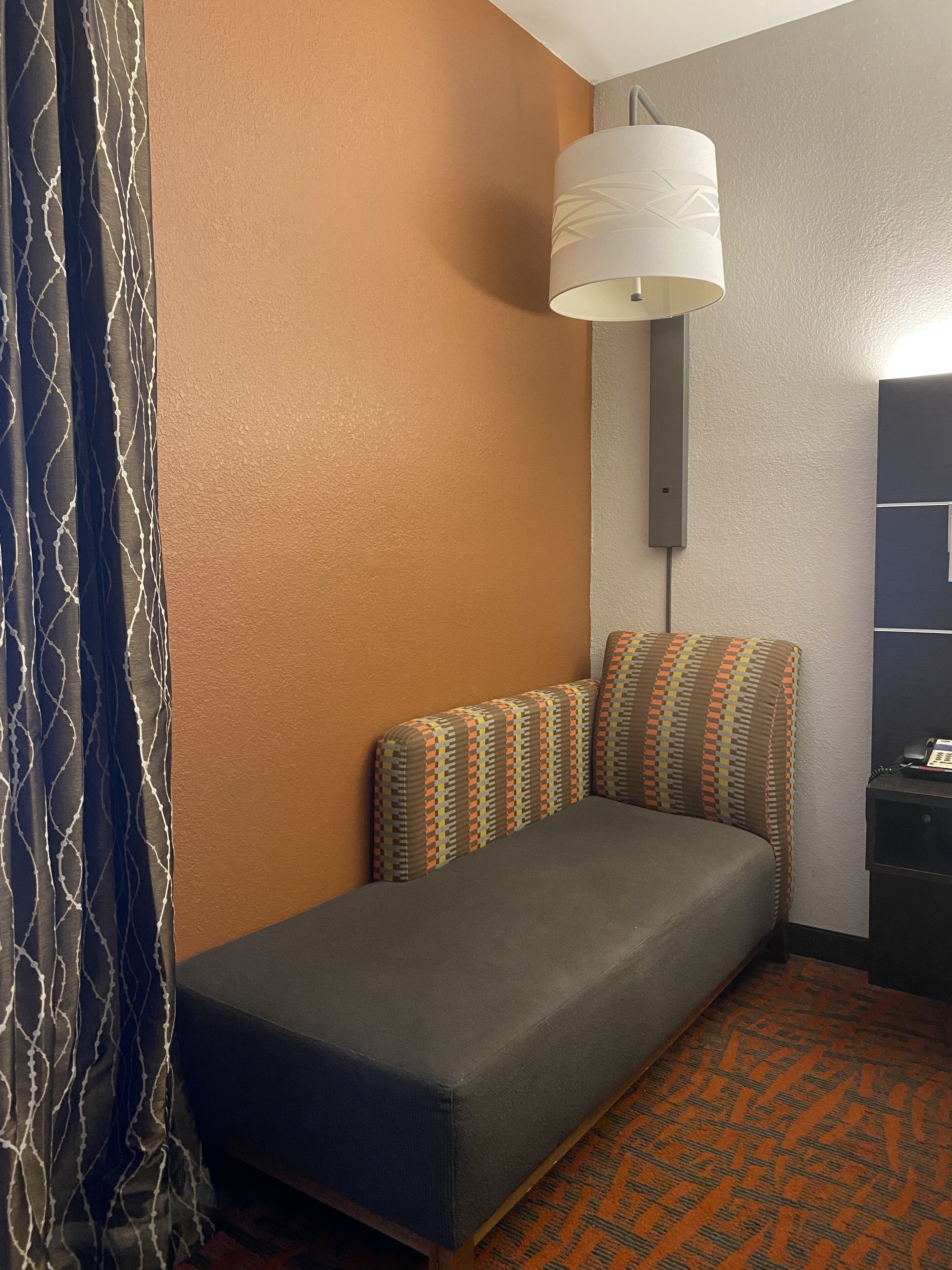 A single bed with patterned pillows, a wall-mounted lamp, and a telephone on a desk.