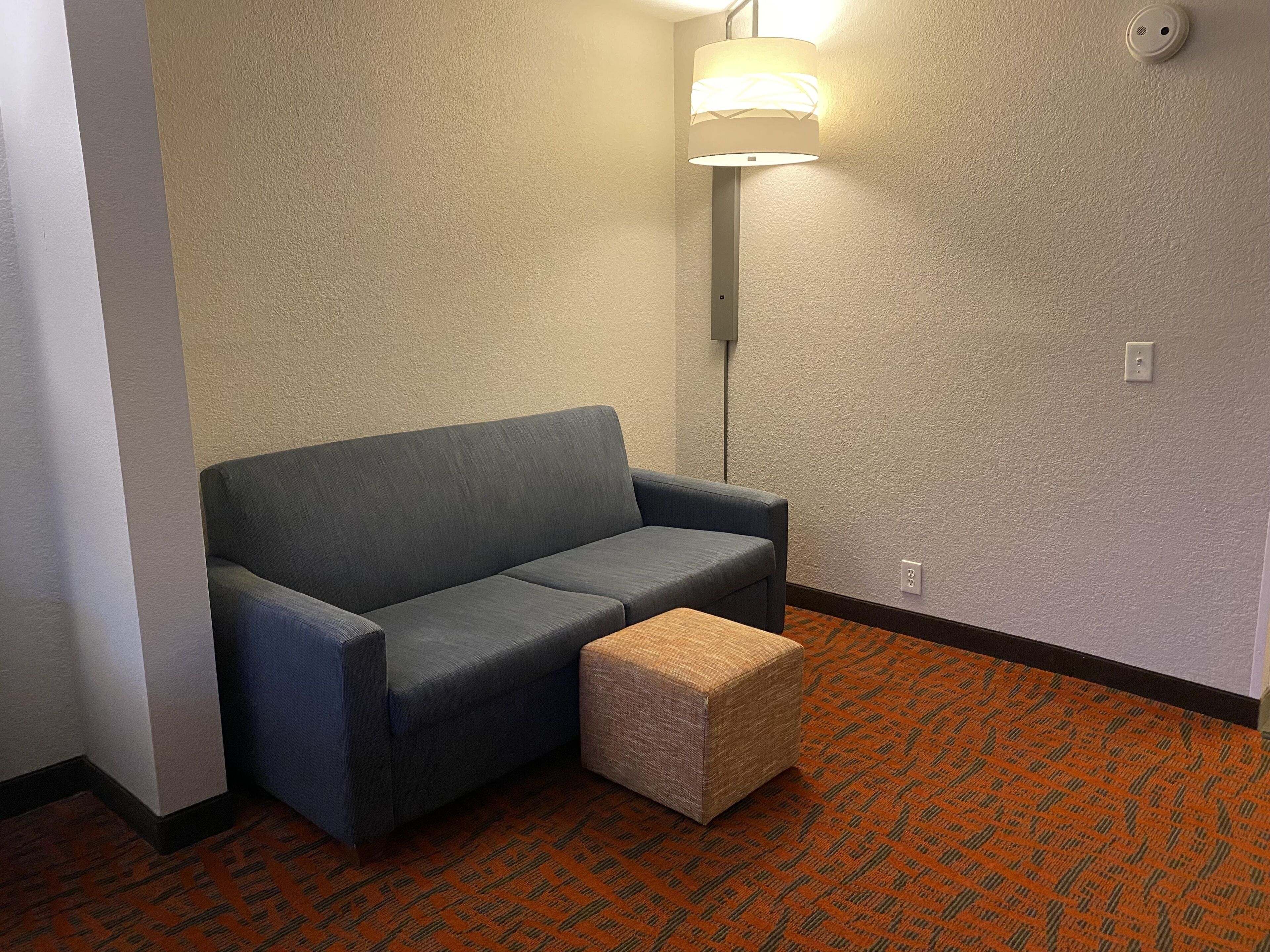 Une pièce d’angle avec un canapé gris, un petit pouf, une lampe fixée au mur et un tapis à motifs.