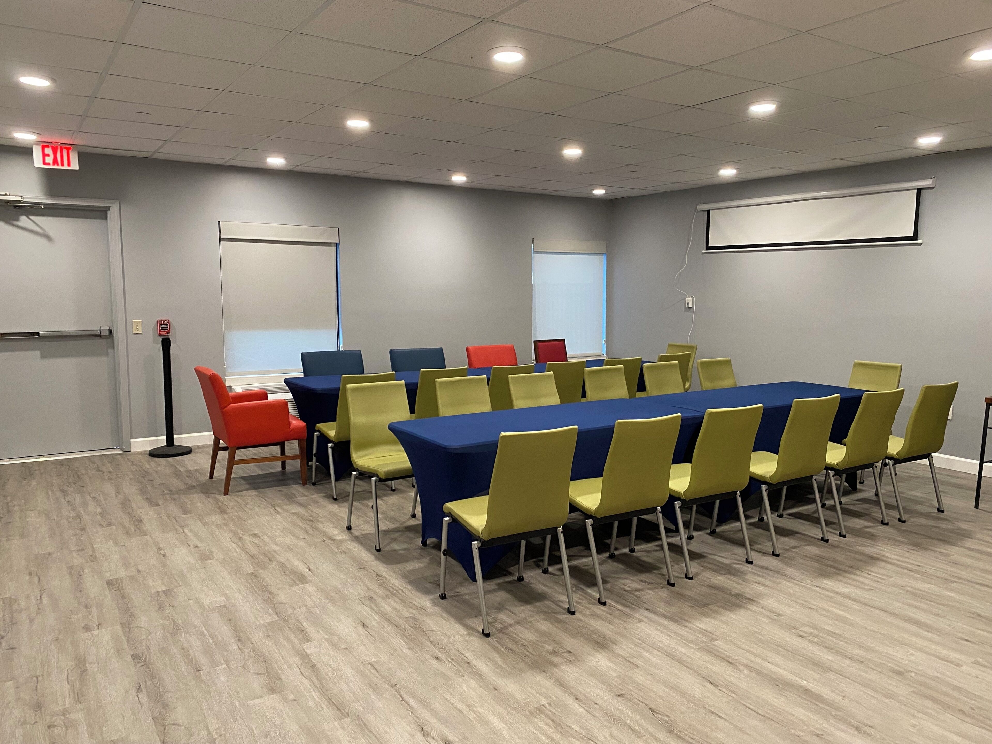 A conference room with a long table, yellow and blue chairs, and a projector screen.