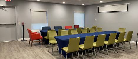 A conference room with a long table, yellow and blue chairs, and a projector screen.