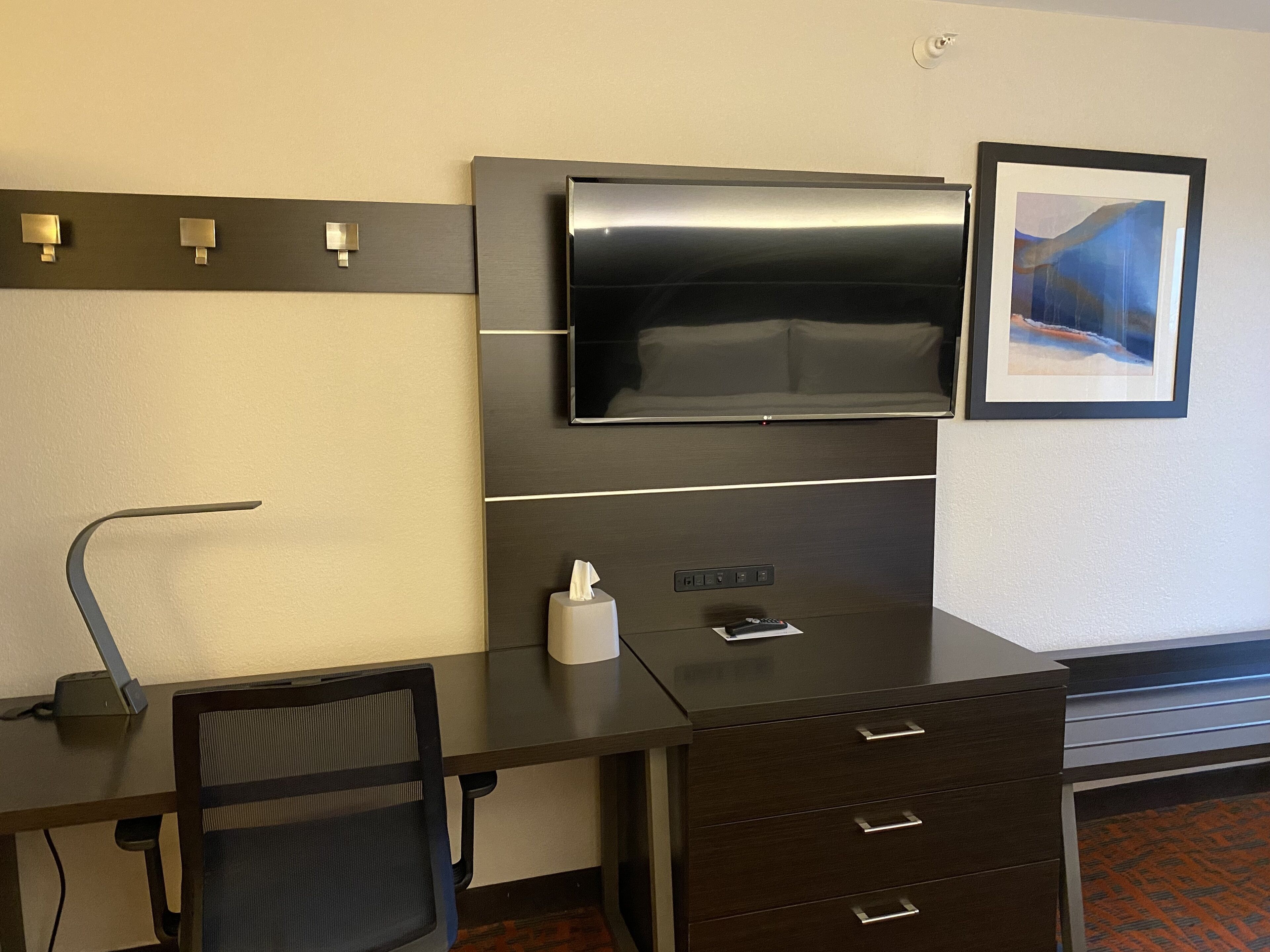 A modern hotel room with a dark wooden desk, a black mesh chair, a flat-screen TV mounted on the wall, a framed picture, and a small bedside table with a remote control and tissues.
