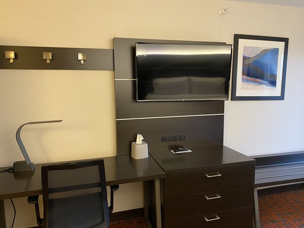 A modern hotel room with a dark wooden desk, a black mesh chair, a flat-screen TV mounted on the wall, a framed picture, and a small bedside table with a remote control and tissues.