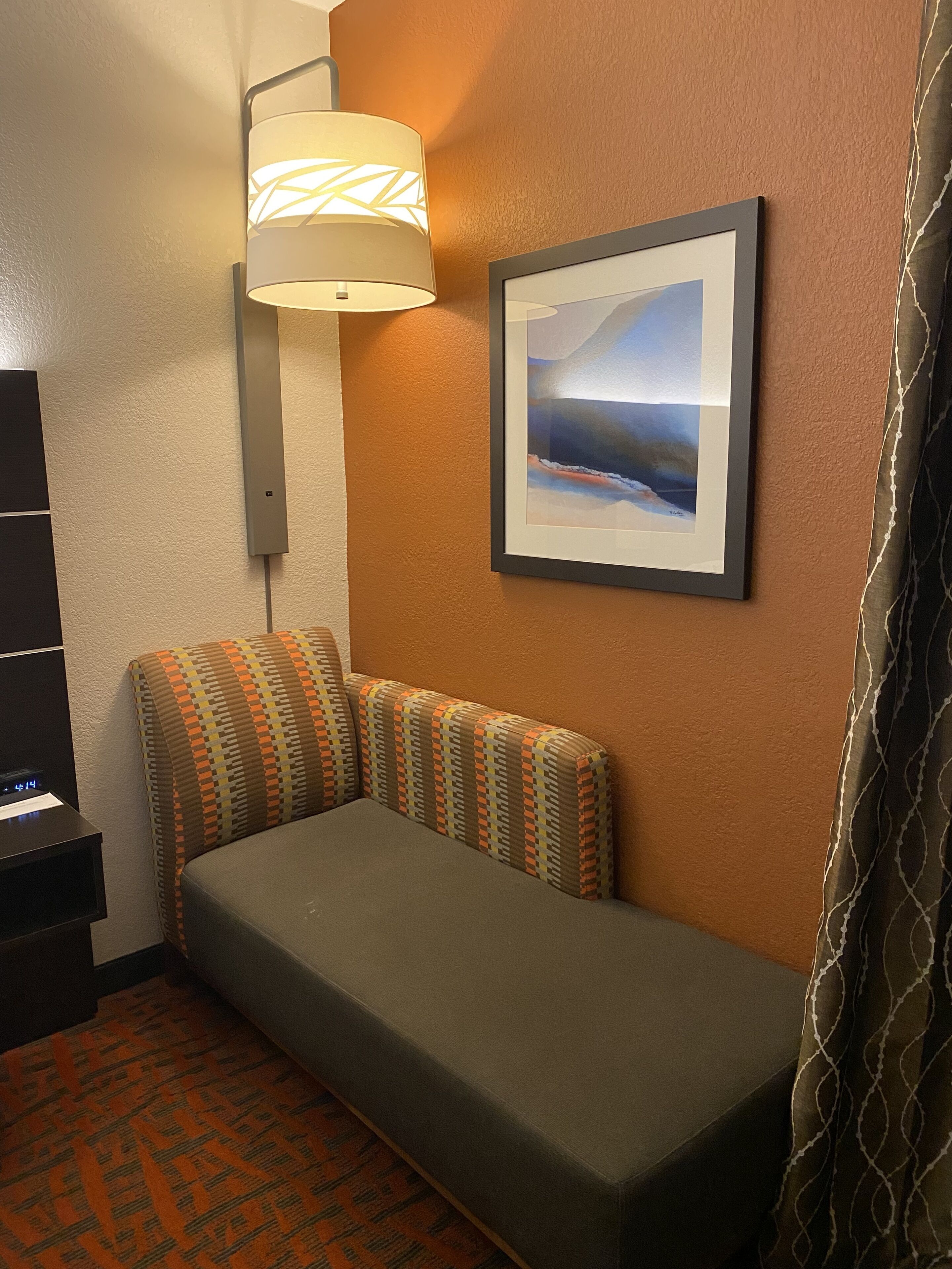 A hotel room with a bed, a wall-mounted lamp, a framed picture, and a digital clock on a nightstand.