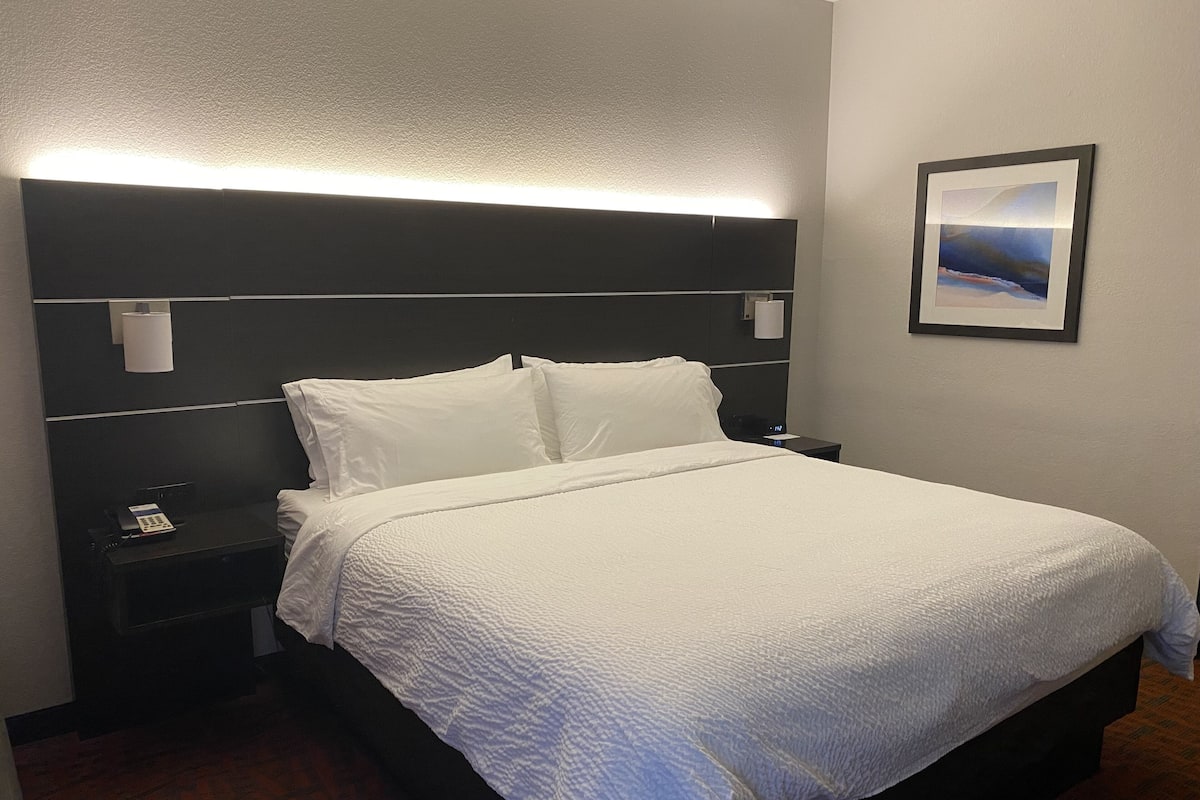 A hotel room with a large bed, a headboard with built-in lighting, a bedside table with a phone, and a framed picture on the wall.