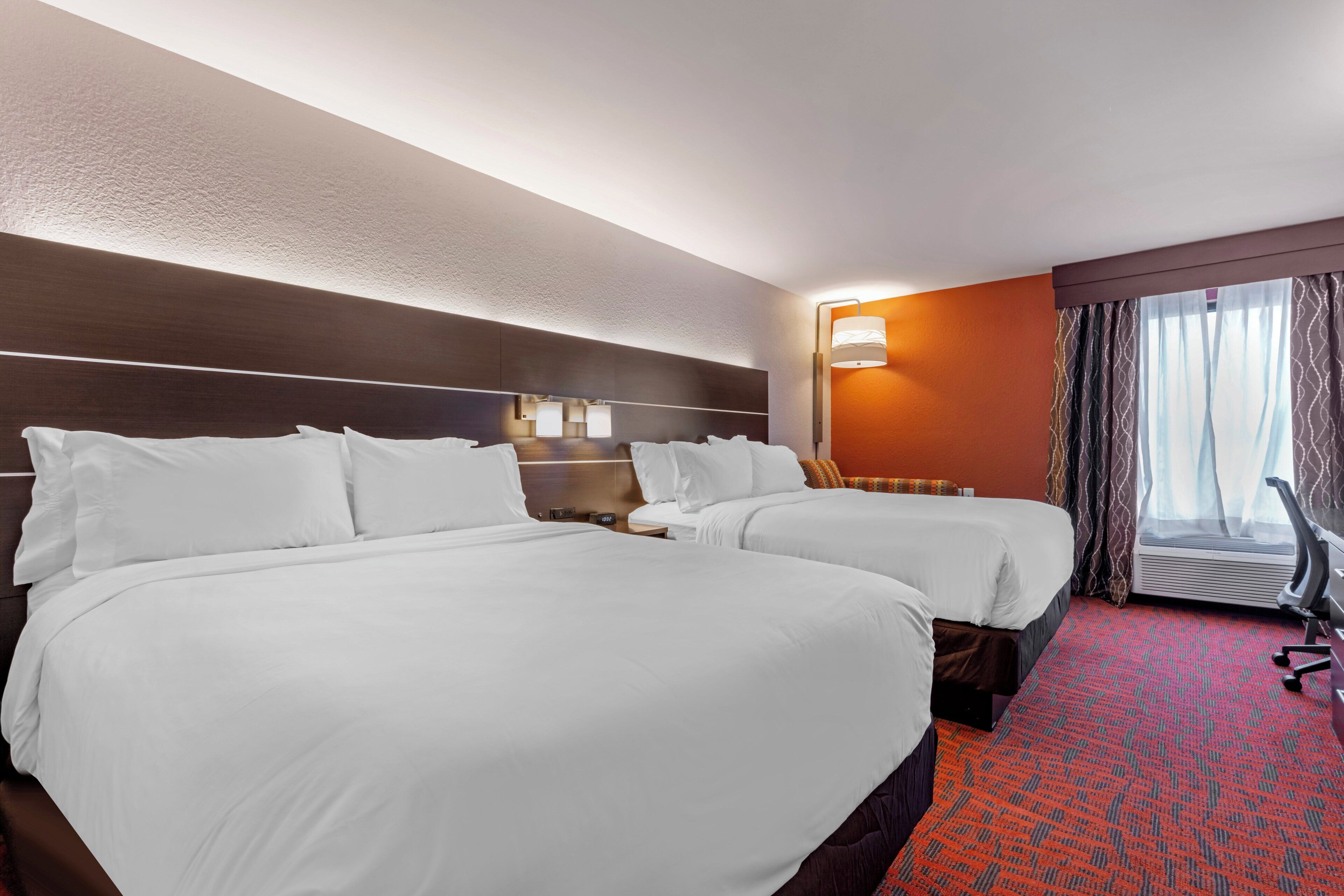 A hotel room with two beds, a desk, a chair, a lamp, and a window with curtains.