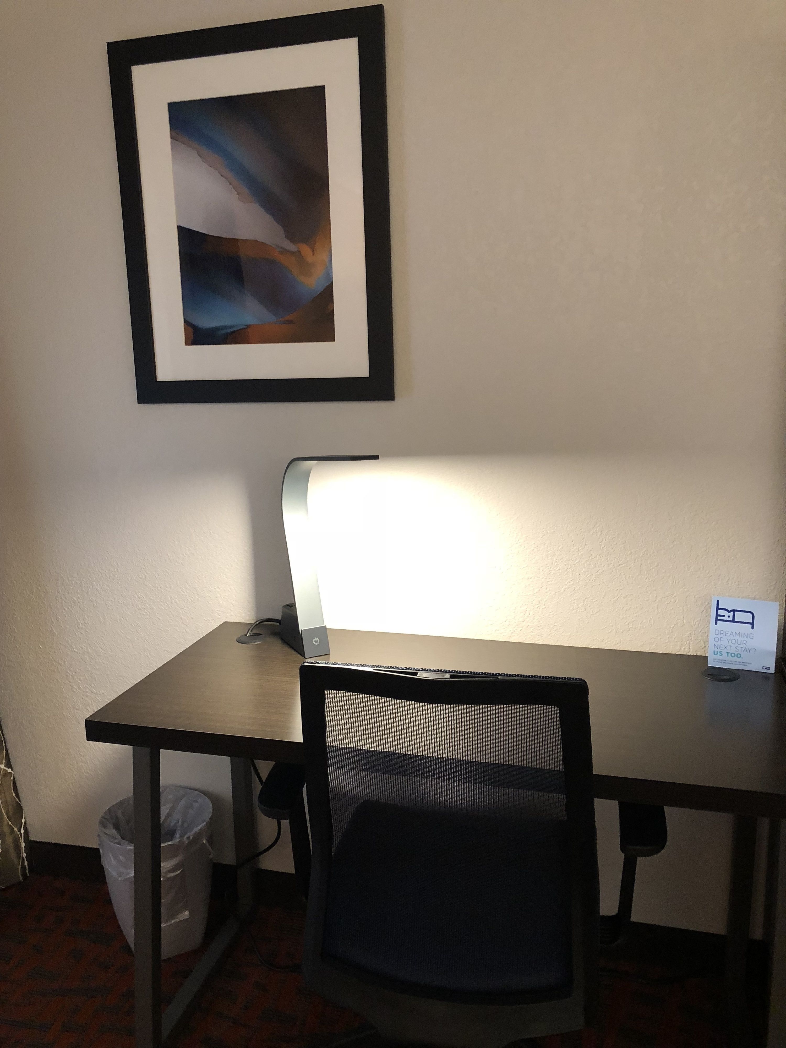 A hotel room with a desk, chair, and a framed abstract painting on the wall.