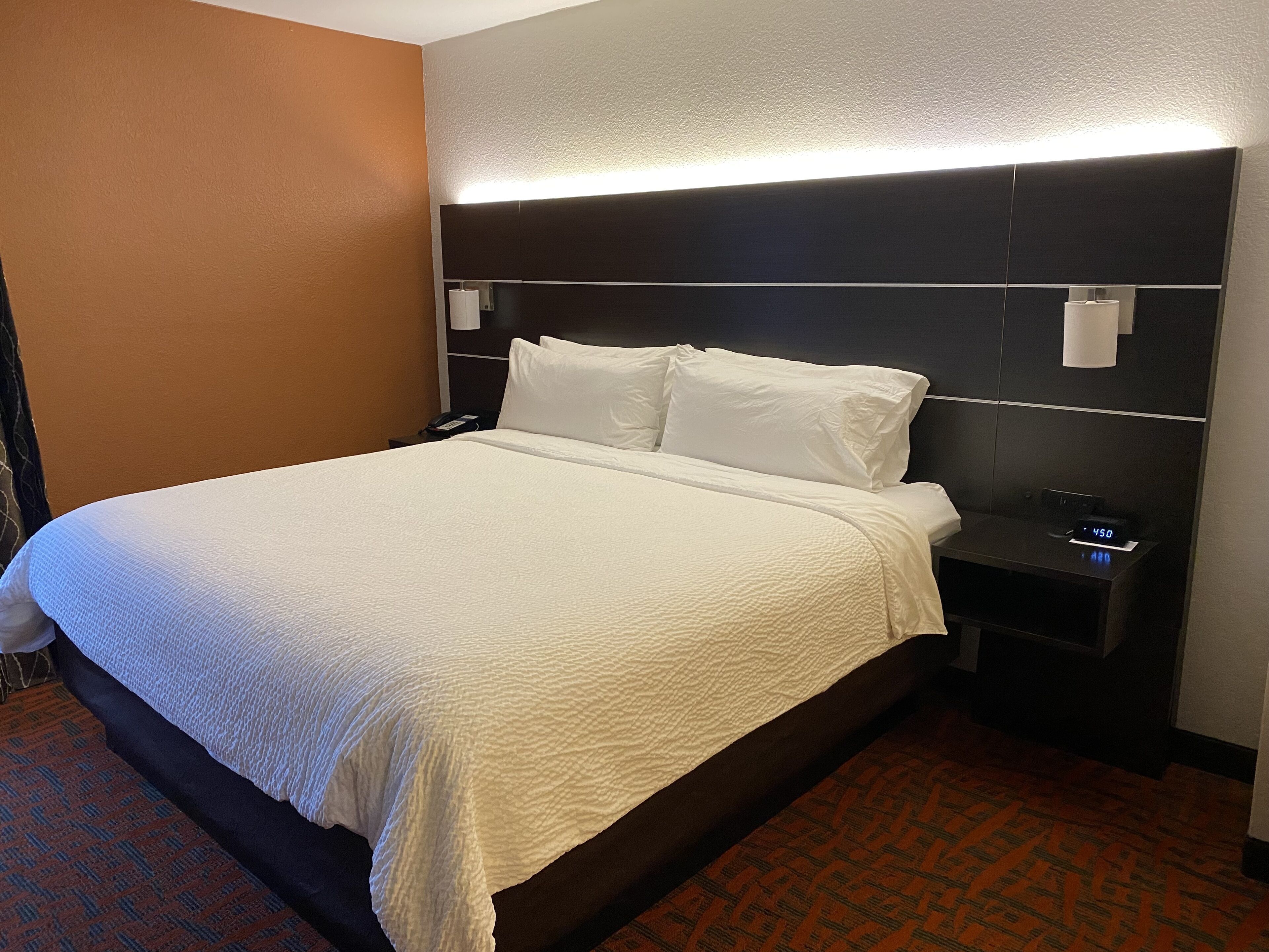 A hotel room with a large bed, a bedside table with a lamp, and a digital clock displaying 4:50.