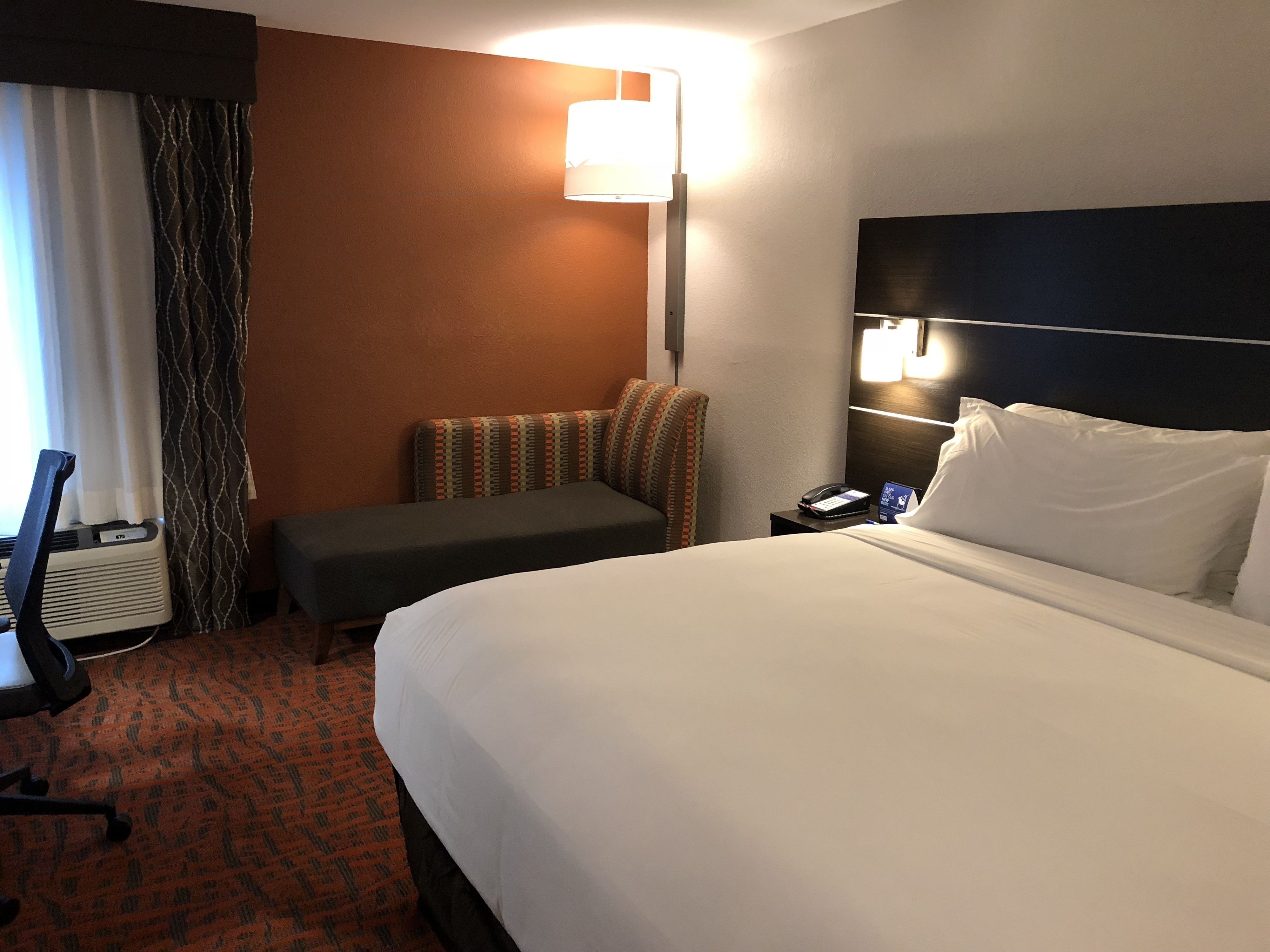 A hotel room with a large bed, a desk with a chair, a small sofa, a bedside table with a lamp, and a window with curtains.