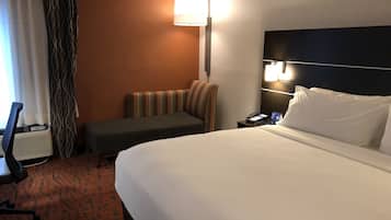 A hotel room with a large bed, a desk with a chair, a small sofa, a bedside table with a lamp, and a window with curtains.
