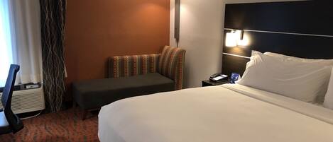 A hotel room with a large bed, a desk with a chair, a small sofa, a bedside table with a lamp, and a window with curtains.