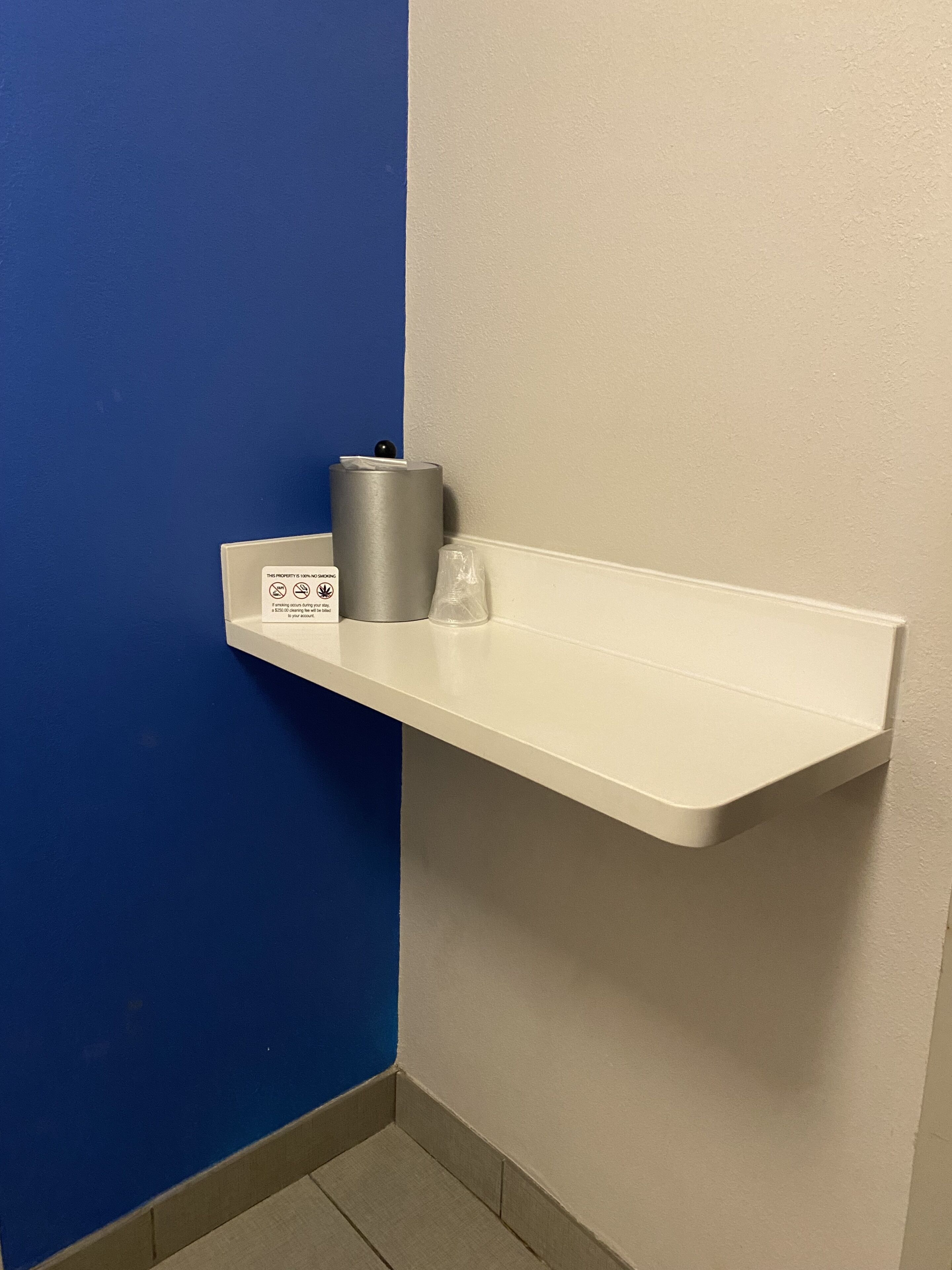 Un estante blanco con un bote de basura y un vaso de plástico frente a una pared azul y blanca.