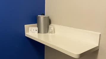 Un estante blanco con un bote de basura y un vaso de plástico frente a una pared azul y blanca.