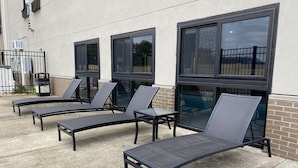 Outdoor lounge chairs and tables arranged on a concrete patio with large windows.