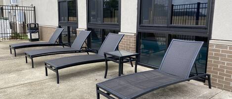 Outdoor lounge chairs and tables arranged on a concrete patio with large windows.