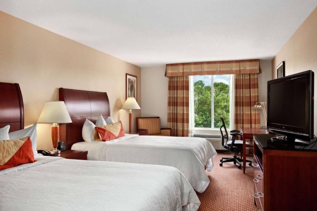 A hotel room with two beds, a television, a desk with a computer, and a window with a view of trees.
