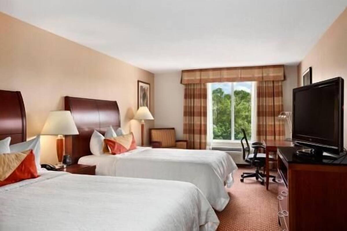 A hotel room with two beds, a television, a desk with a chair, and a window with a view of trees.