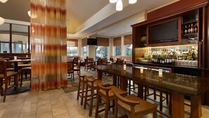 A bar area with a long counter, multiple stools, and a television screen above the bar. - Hilton Garden Inn Chicago / Oak Brook (Villa Park)