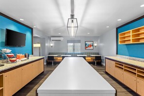 A modern hotel room with a long white table, seating, a flat-screen TV, and a buffet setup. - MainStay Suites Chicago Schaumburg (Schaumburg)