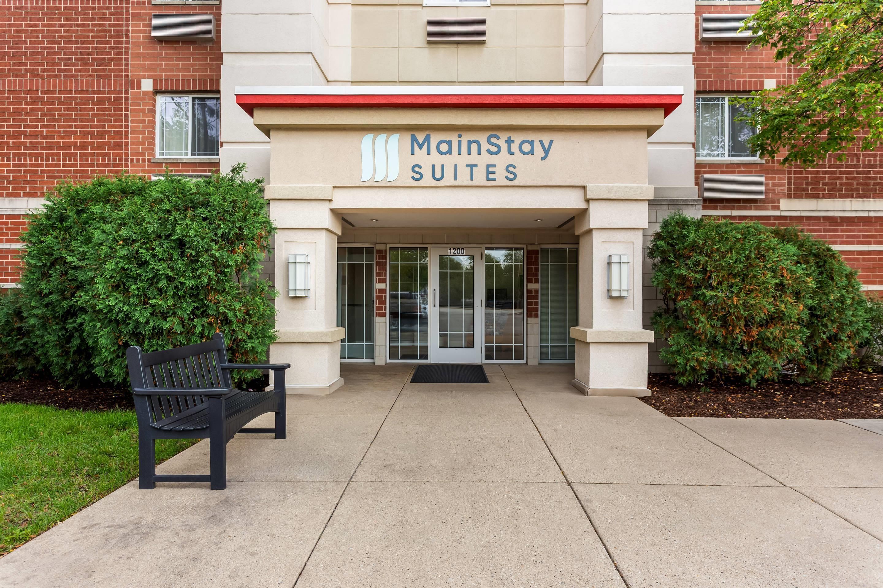 Entrée de l’hôtel MainStay Suites, avec une façade en briques, des portes en verre et un banc à l’extérieur.