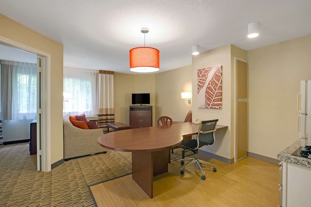 A hotel room with a wooden dining table, a sofa, a TV, and a kitchen area.