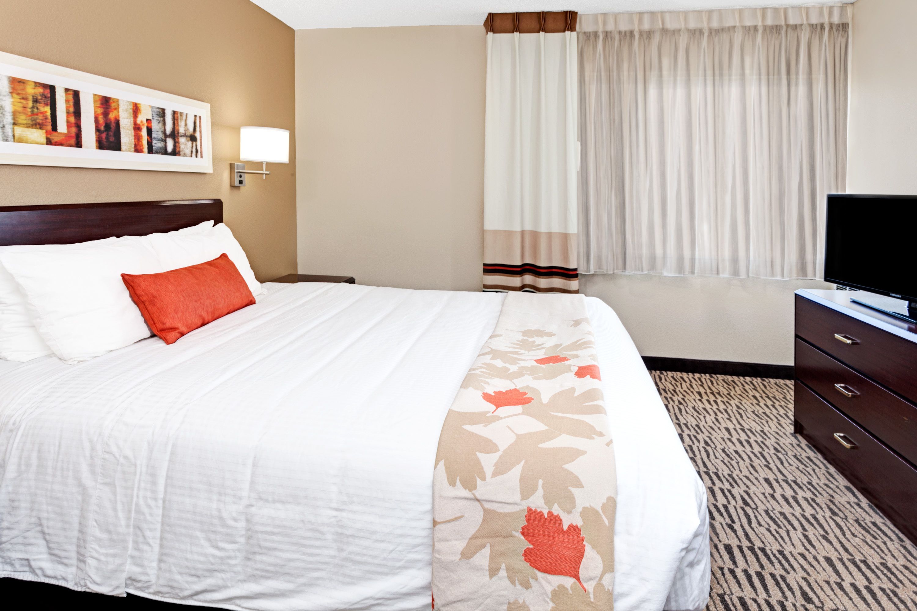 A hotel room with a large bed, a flat-screen TV, a dresser, and a window with curtains.