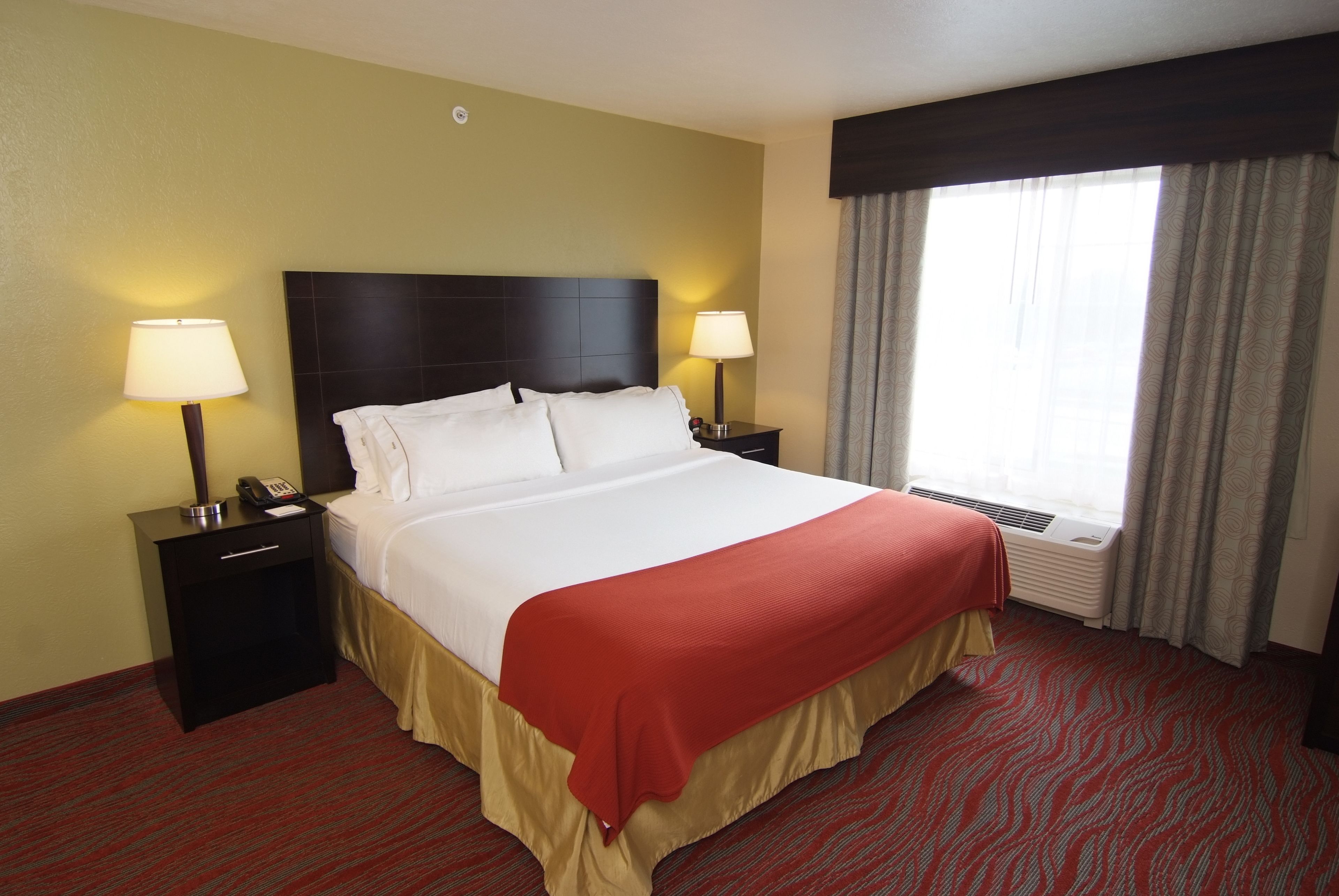 A hotel room with a large bed, two bedside tables with lamps, a window with curtains, and a red carpet.
