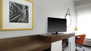A modern hotel room with a wooden desk, a flat-screen TV, a framed black and white photo, and a bright orange chair. - Hampton Inn Jasper (Jasper)