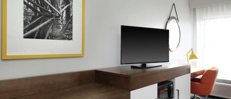 Ett modernt hotellrum med ett skrivbord i trä, en platt-TV, ett inramat svartvitt foto och en knallorange stol.