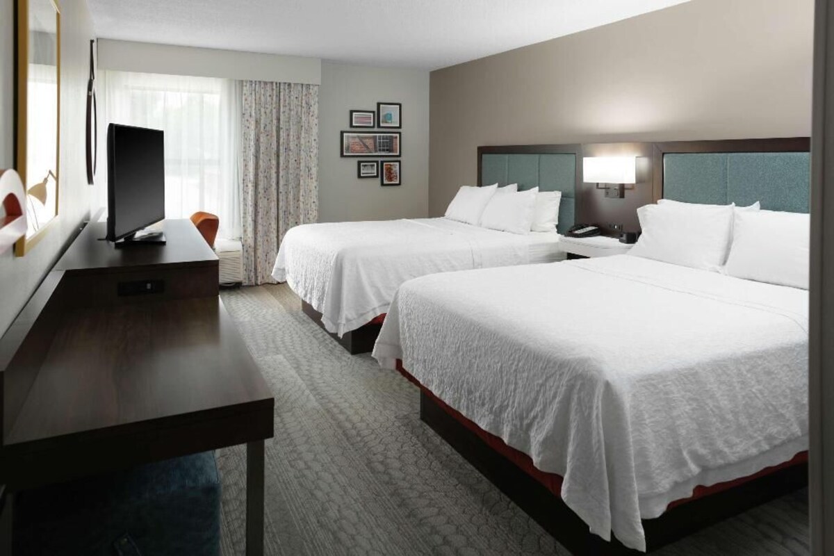 Quarto de hotel com duas camas, uma escrivaninha, uma televisão e janela com cortinas.