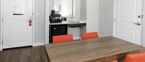 Habitación de hotel con una mesa de comedor de madera, sillas anaranjadas, una zona de cocina con microondas y un lavabo.