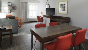 A modern hotel room with a wooden dining table, red chairs, a grey sofa, a small desk with a lamp, a flat-screen TV, and framed pictures on the wall. - Hampton Inn Jasper (Jasper)