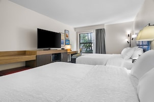 A hotel room with two beds, a flat-screen TV, a desk with a lamp, and a window with curtains.