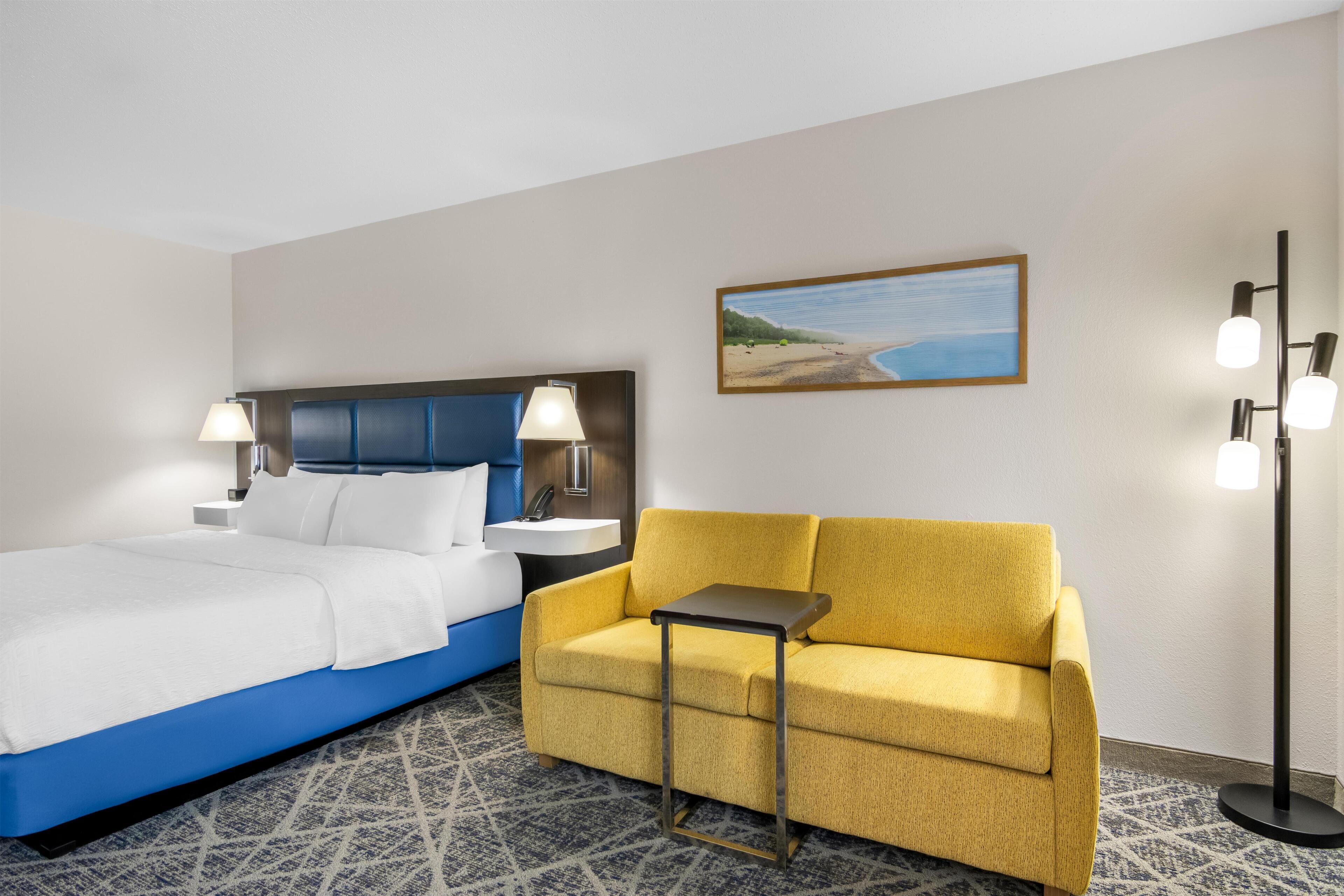 Una habitación de hotel con cama, sofá, mesita de noche, lámpara, un cuadro en la pared y una lámpara de pie.