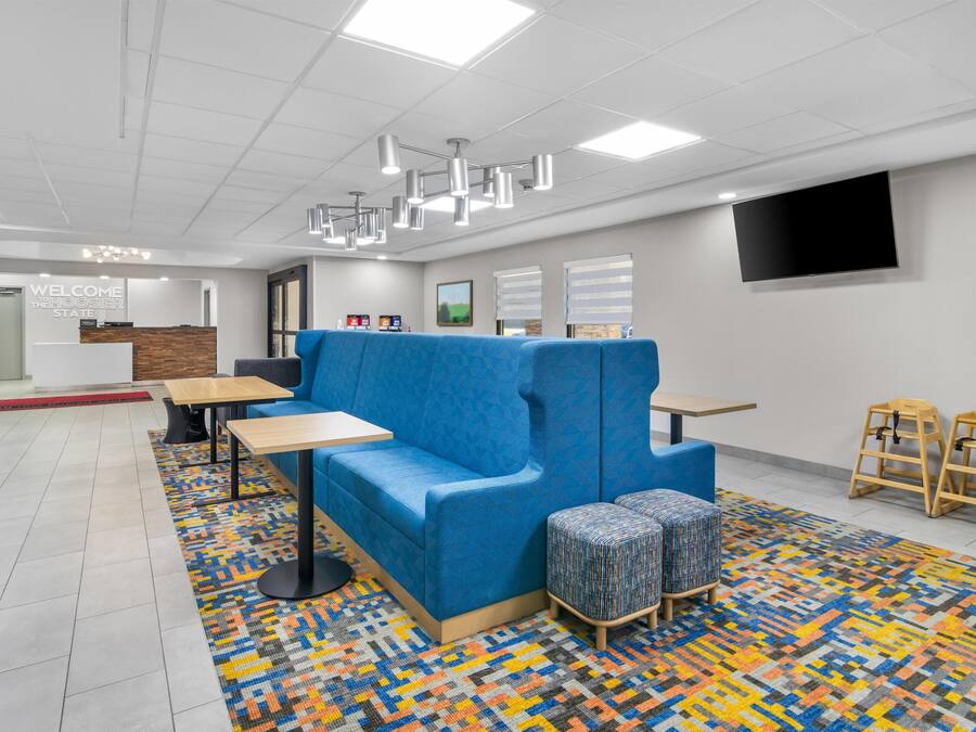 A modern hotel lobby with a blue sofa, wooden tables, a patterned rug, and a flat-screen TV on the wall.