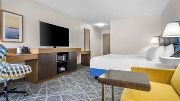 A hotel room with a bed, a desk with a flat-screen TV, a chair, a small table, and a lamp.
