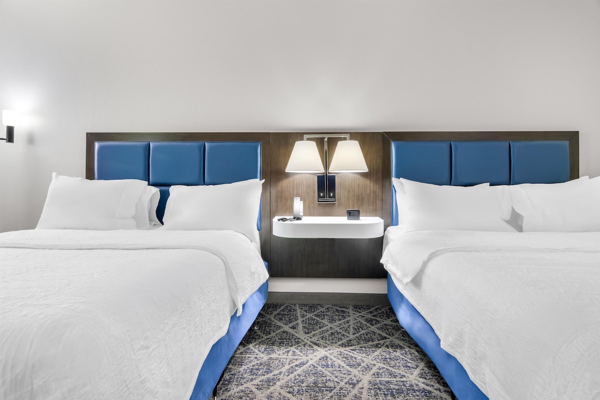 A hotel room with two beds, a nightstand with a lamp, and a small sink.