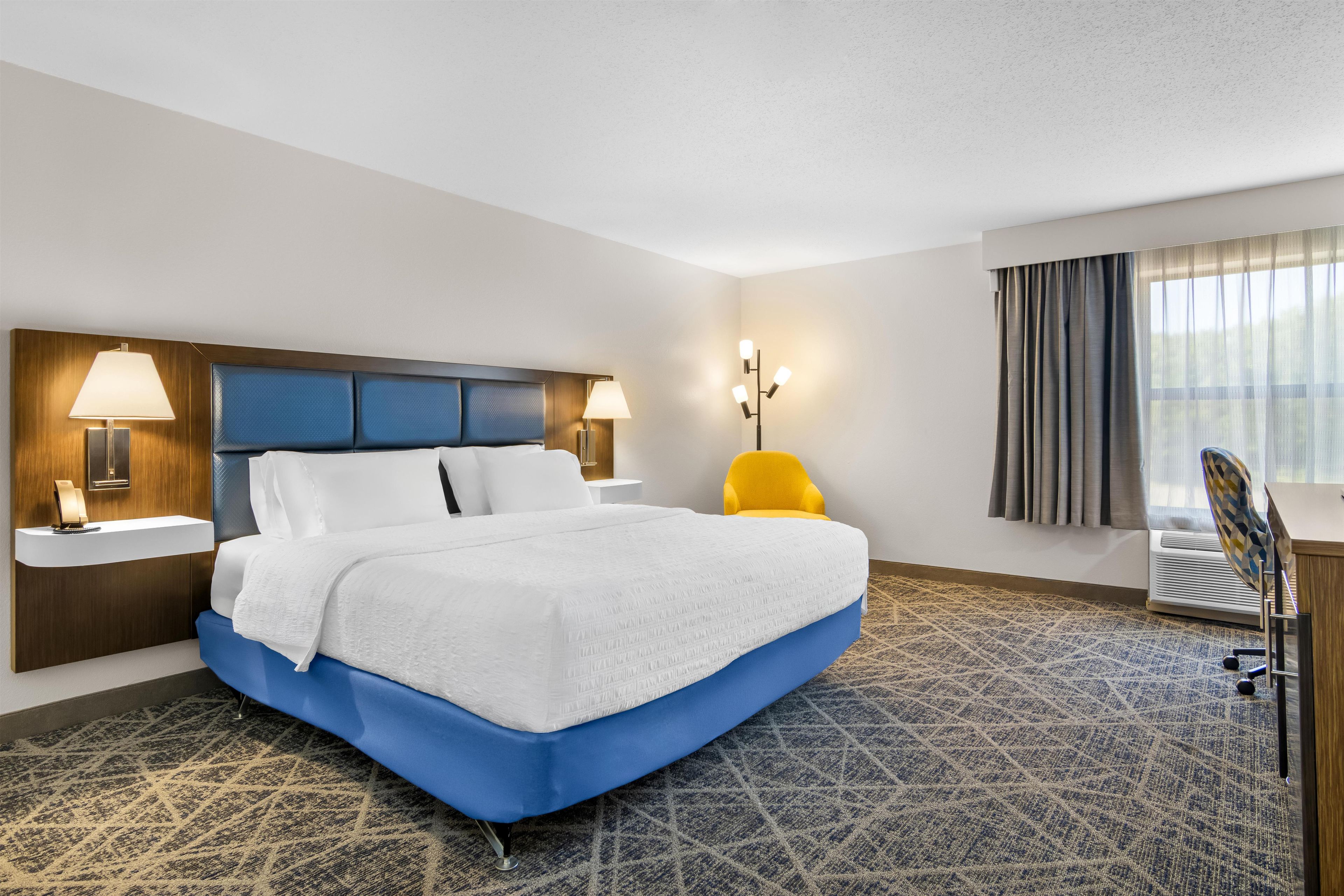 Quarto de hotel com uma cama grande, uma escrivaninha, uma cadeira e uma janela com cortinas.