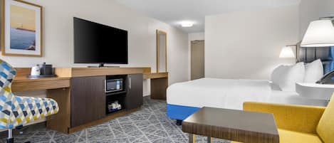 A hotel room with a bed, a desk with a flat-screen TV, a chair, a small table, and a lamp.