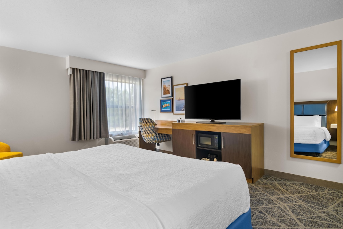 A hotel room with a bed, a desk with a flat-screen TV, a chair, a microwave, and a large mirror.