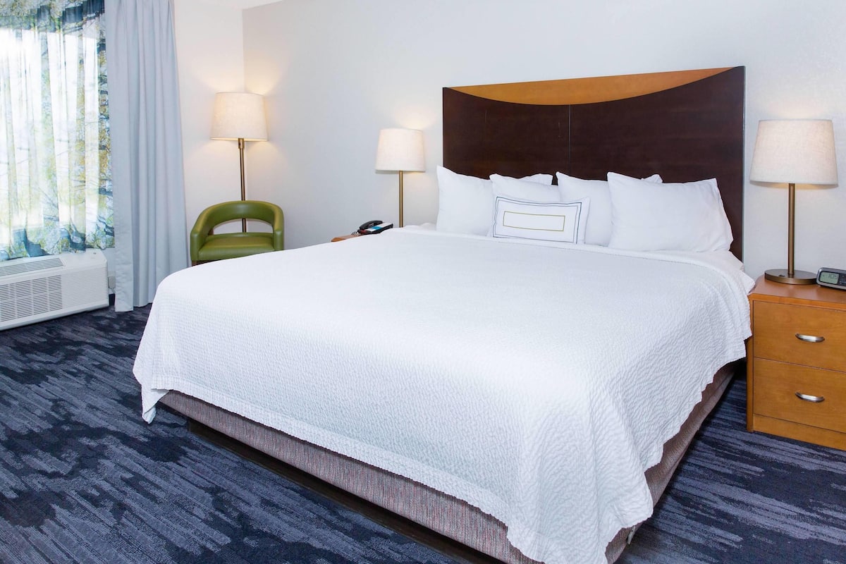 A hotel room with a large bed, two bedside lamps, a green chair, a bedside table with a phone, and a window with curtains.