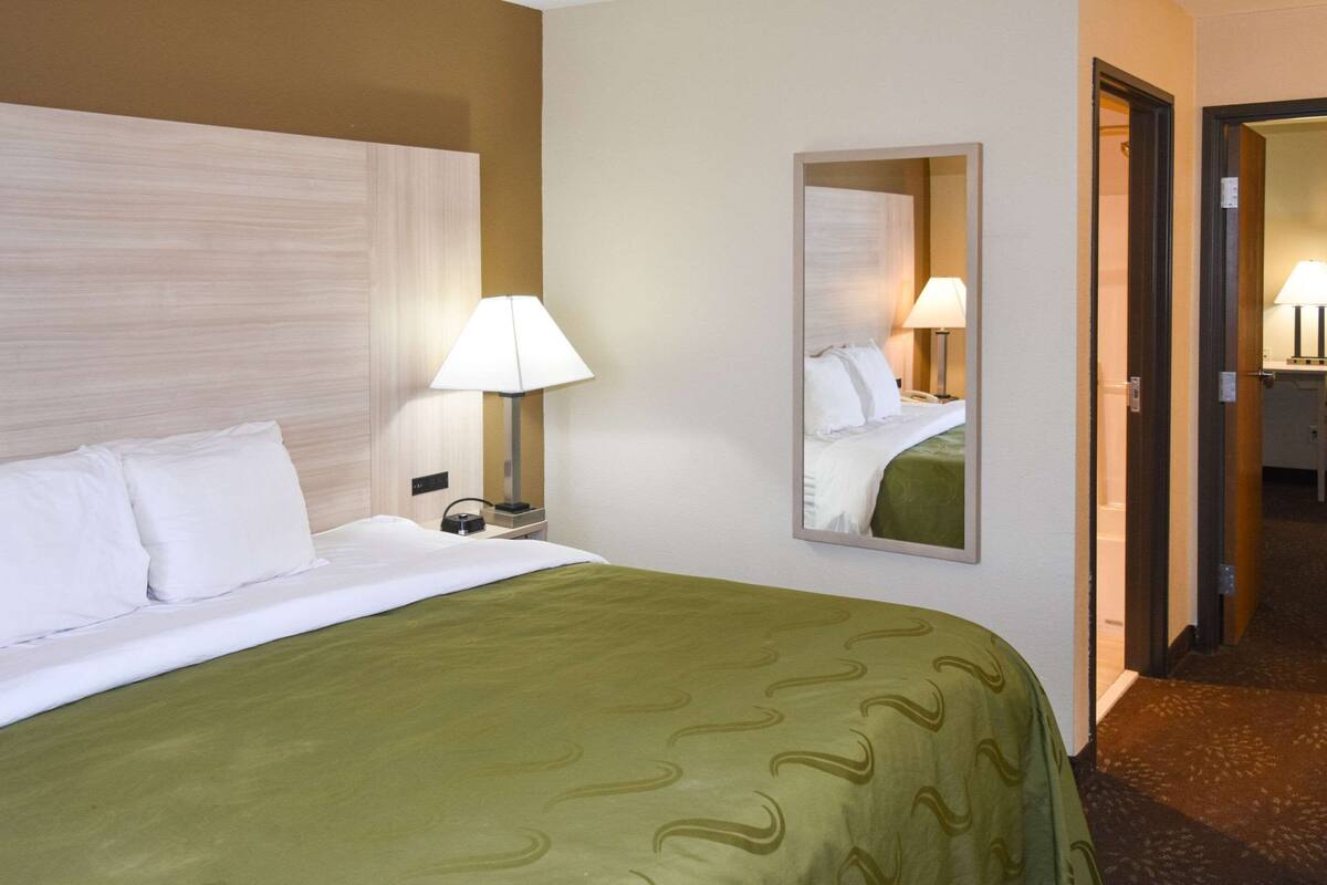 Quarto de hotel com uma cama grande, abajur, espelho e porta para um banheiro.