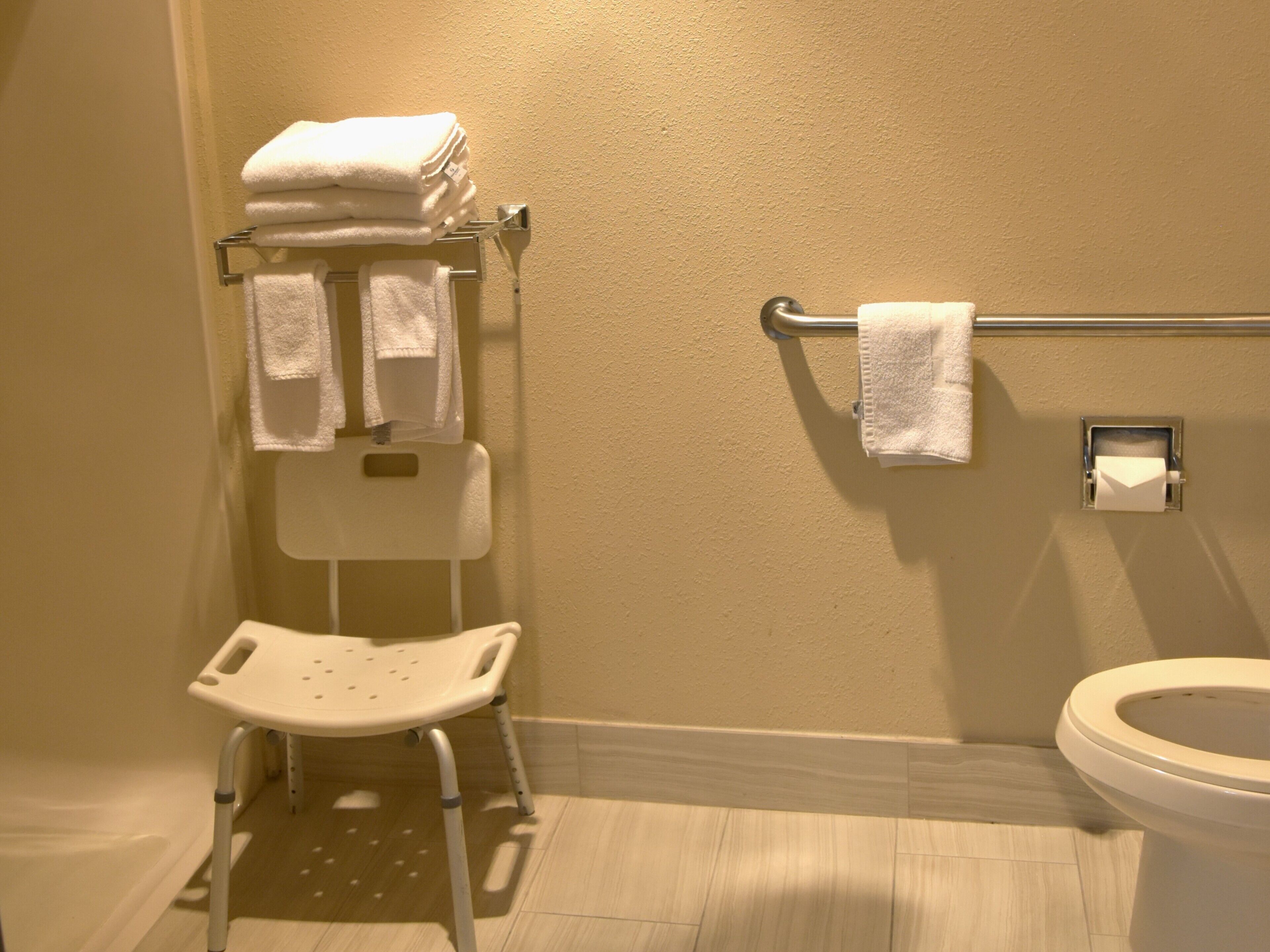 A bathroom with a white toilet, a white sink, a white shower chair, and a towel rack with folded towels.
